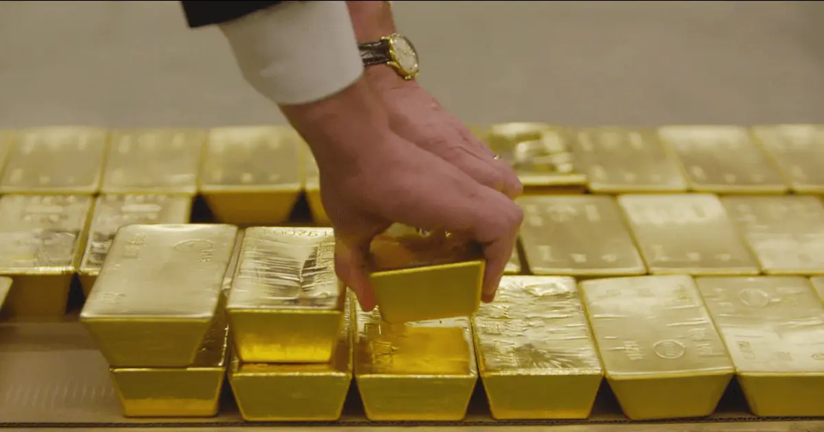 A person's hand reaching down to pick up a gold bullion bar from a large collection of stacked bars.