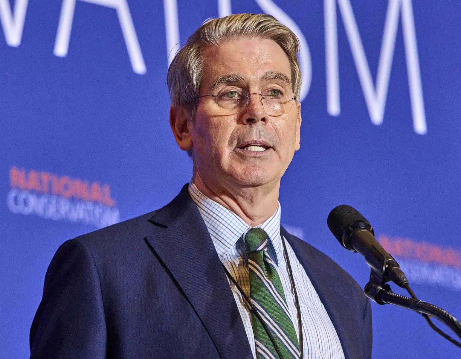 Scott Bessent speaking at a podium with a blue background and a microphone in front of him.