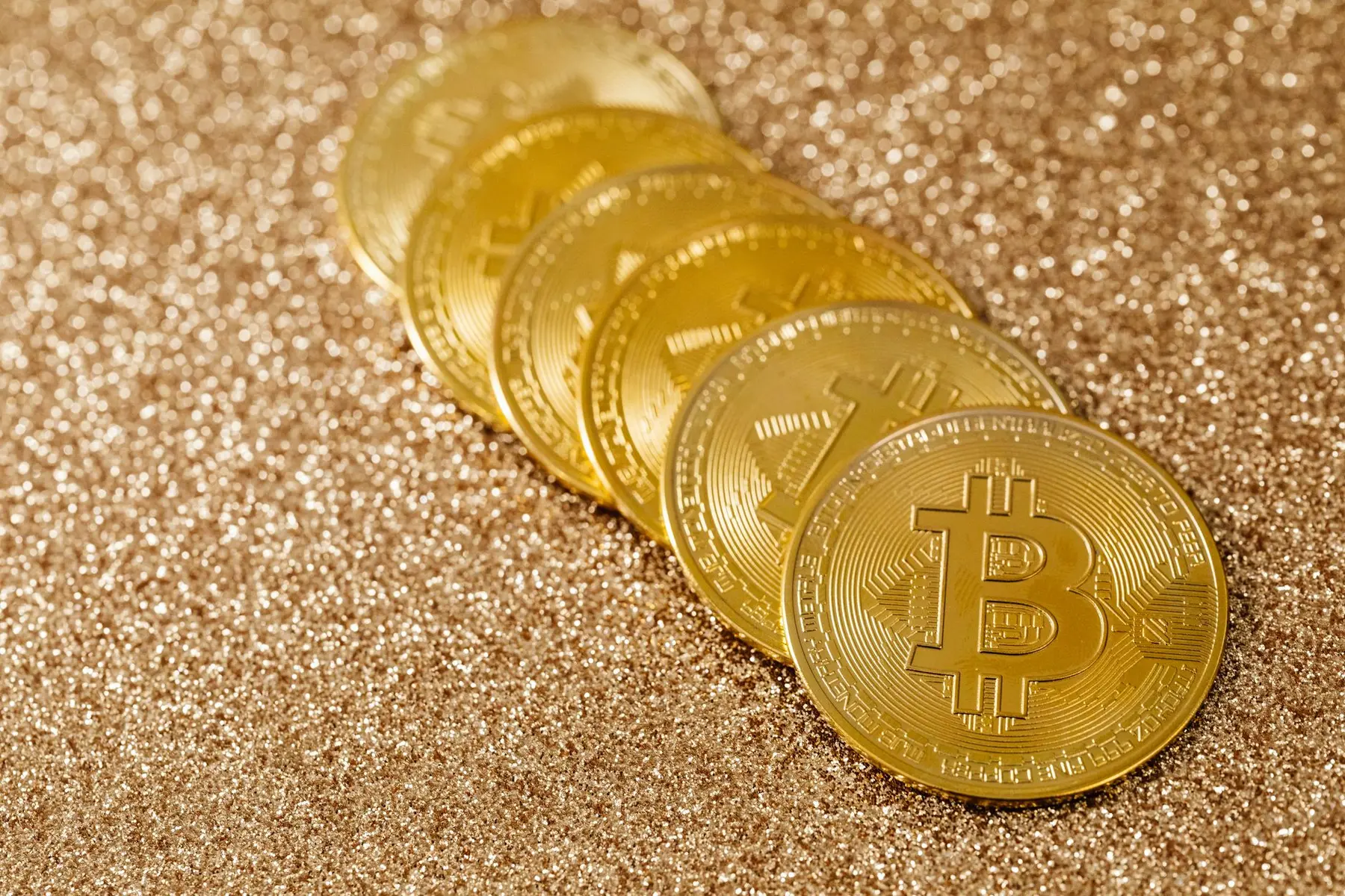 A row of gold-colored physical Bitcoin coins resting on a shimmering gold glitter background
