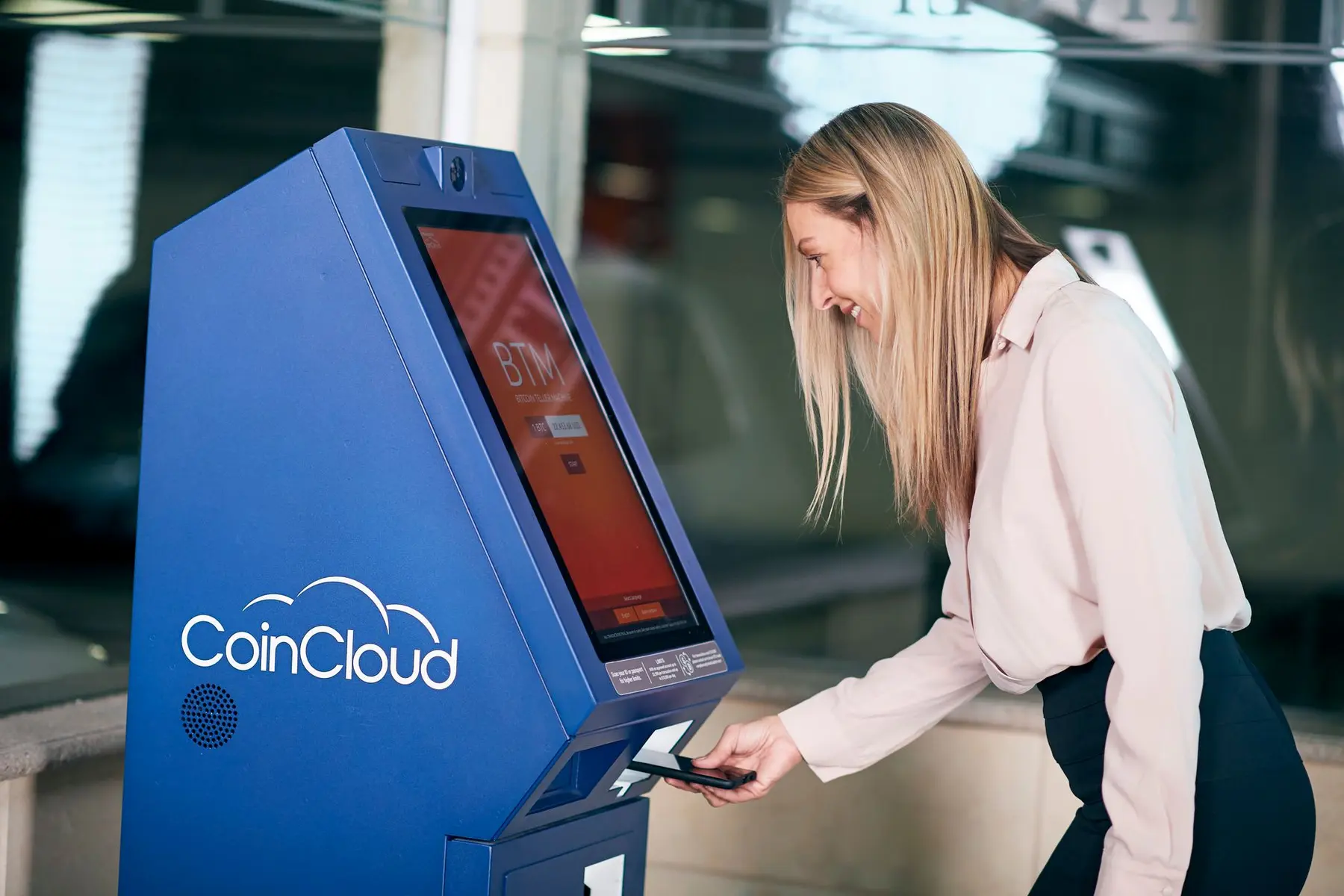 A woman using a smartphone to make a contactless payment at a blue CoinCloud Bitcoin ATM