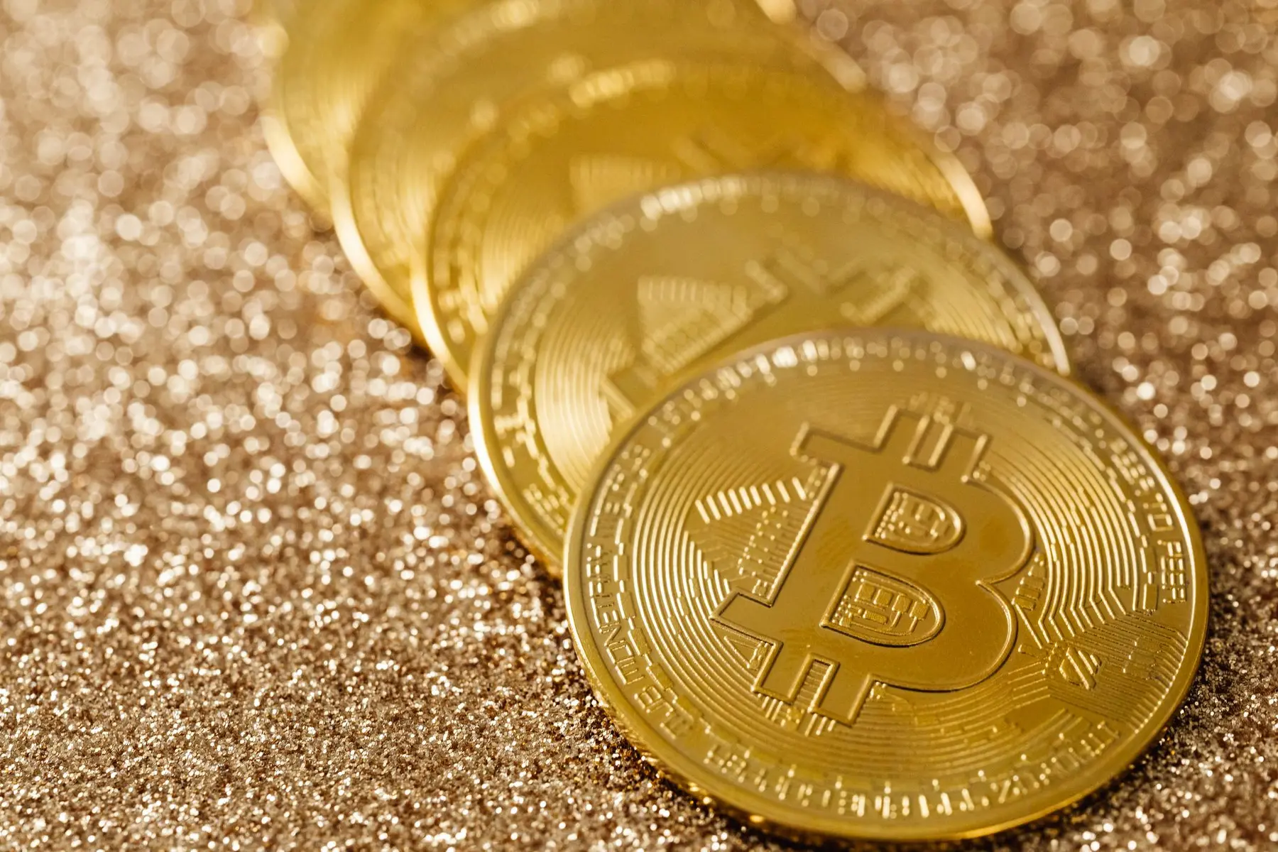 A row of gold Bitcoin coins resting on a shimmering gold glitter surface