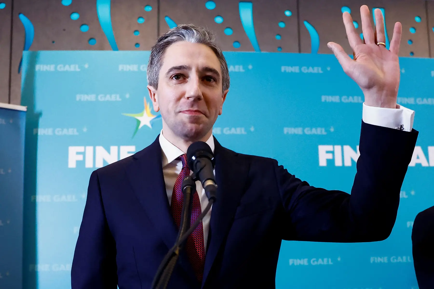 Simon Harris waving his hand while standing behind a microphone in front of a Fine Gael backdrop