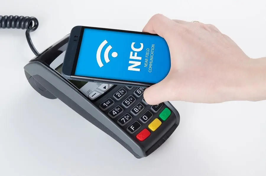 A hand holding a smartphone with an NFC icon over a credit card payment terminal