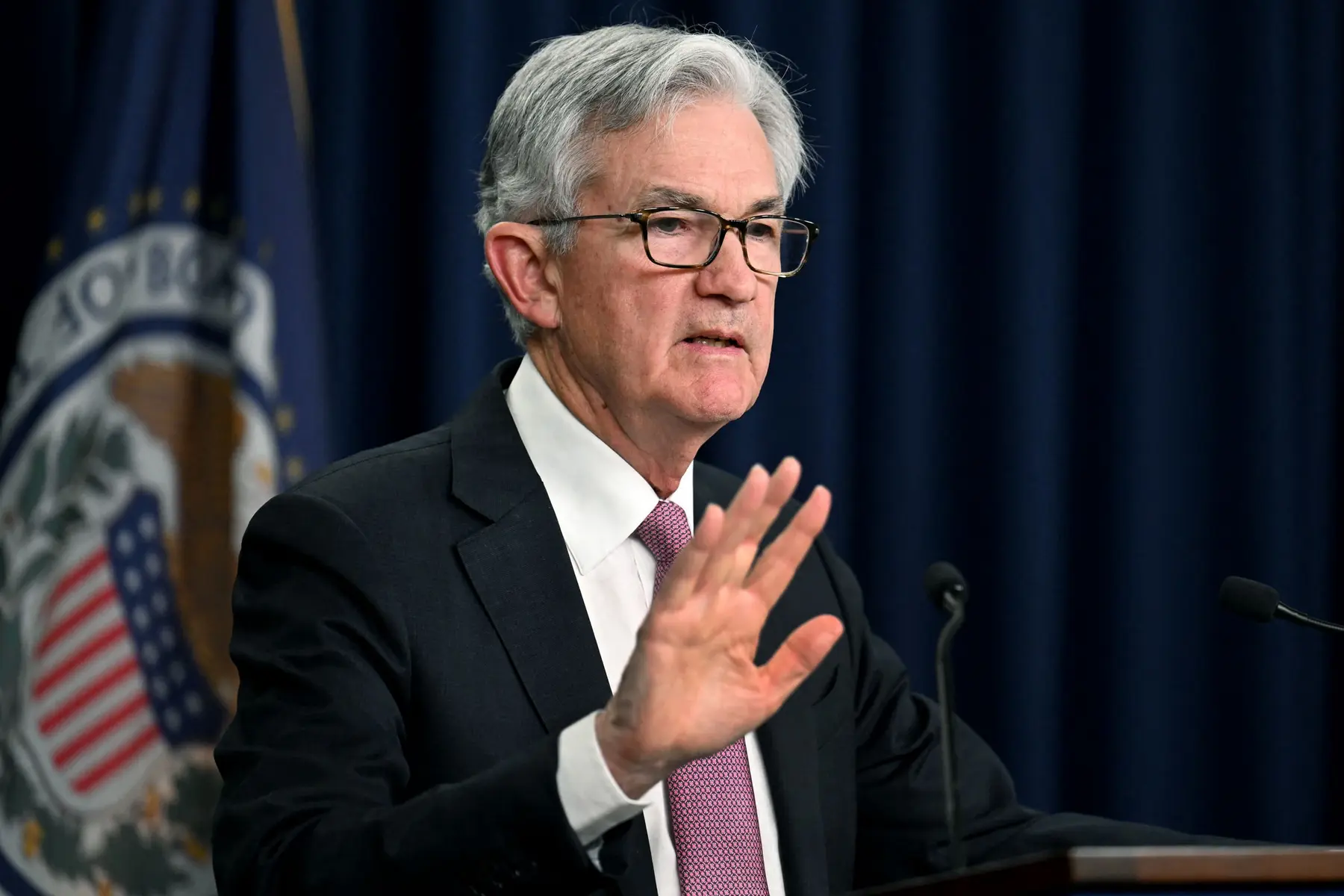 Jerome Powell speaking at a podium with a Federal Reserve seal flag in the background