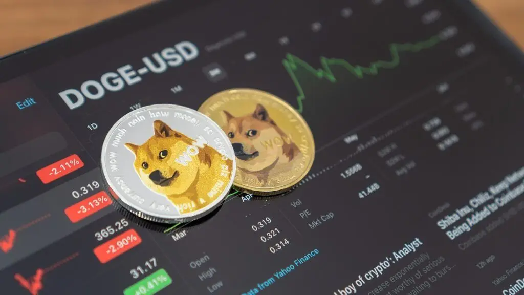 Two physical Dogecoin tokens featuring the Shiba Inu dog resting on a digital screen showing a DOGE-USD price chart.