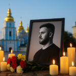A bright candlelit memorial on Kyiv’s cobblestones features a framed black-and-white portrait of Konstantin Galich surrounded by glowing candles and red-yellow flowers, with the sunlit blue-and-gold domes of St. Michael’s softly out of focus behind.