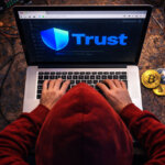 A hacker in a red hoodie uses a laptop displaying the Trust Wallet logo as Bitcoin and Ethereum coins sit nearby, illustrating how browser wallets leaked $713M in 2025.