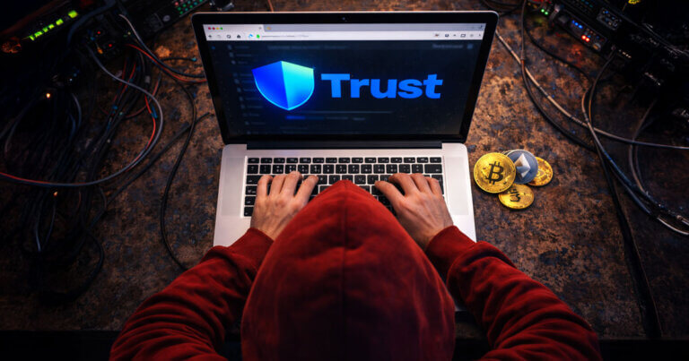 A hacker in a red hoodie uses a laptop displaying the Trust Wallet logo as Bitcoin and Ethereum coins sit nearby, illustrating how browser wallets leaked $713M in 2025.